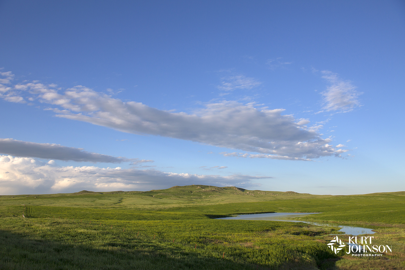Hopeful blue sky with puffy low drifting white clouds above a field of bright green grass with a small river flowing through it is a picturesque scene for healing healthcare environments.