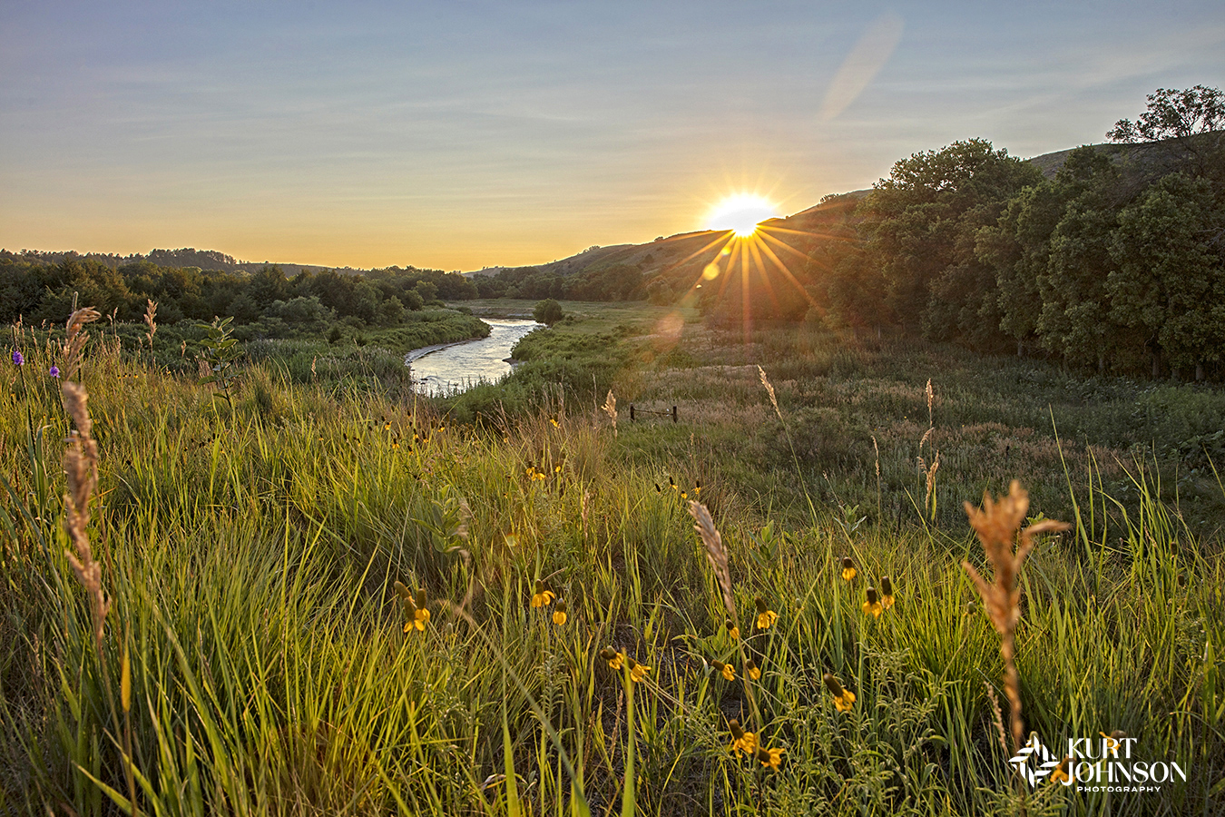 midwest wildflower landscape with winding river at sunset as the sun fades behind a lush hill with sunlight bursting in a star shape across a field of tall grasses